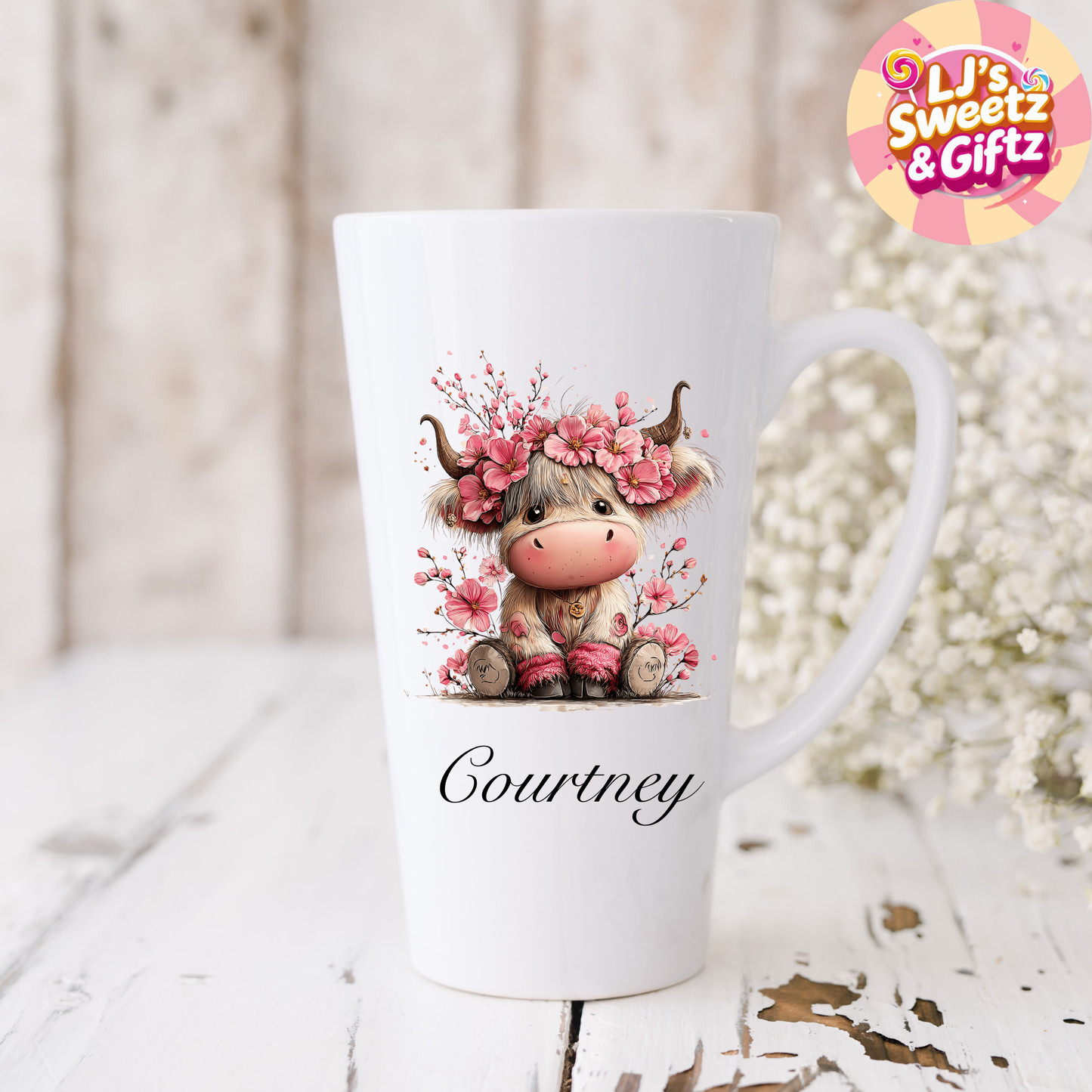 White mug with a cartoon cow wearing a floral headband and the name 'Courtney' on a wooden surface with flowers.