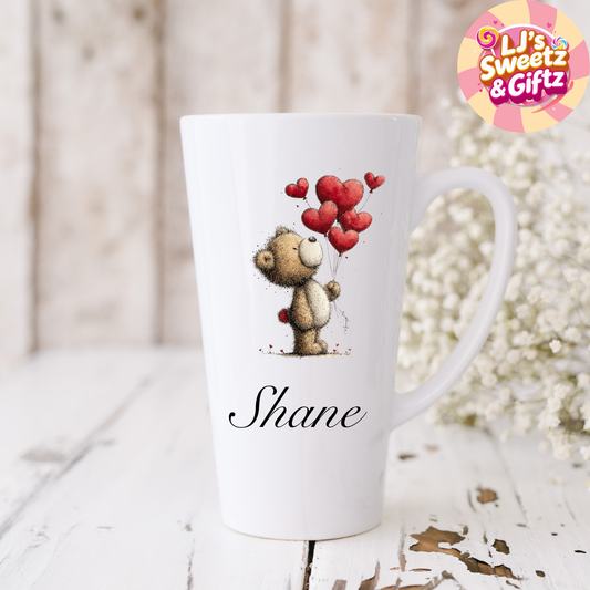 White mug with teddy bear holding heart balloons and 'Shane' printed on it, placed on a wooden surface with decorative elements.