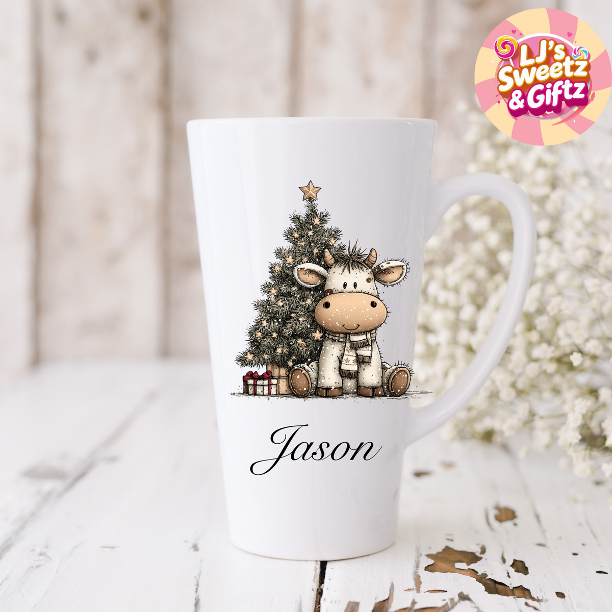 White mug with a cow and Christmas tree design, personalized 'Jason', on a light wooden surface with decorative elements.