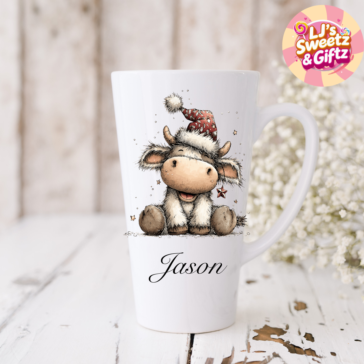 White mug with a cartoon cow wearing a Santa hat and the name 'Jason' on a wooden surface with decorative elements.